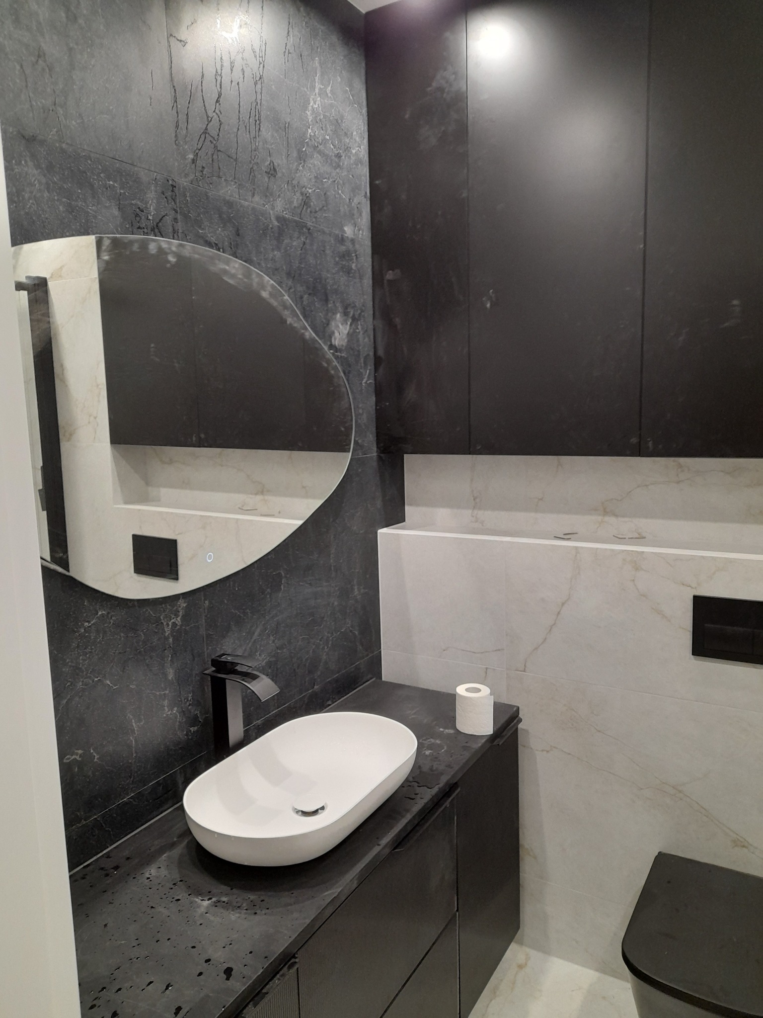Nowoczesna łazienka z umywalką nablatową, czarną armaturą i marmurowymi płytkami. Asymetryczne lustro z podświetleniem LED dodaje unikalnego charakteru.