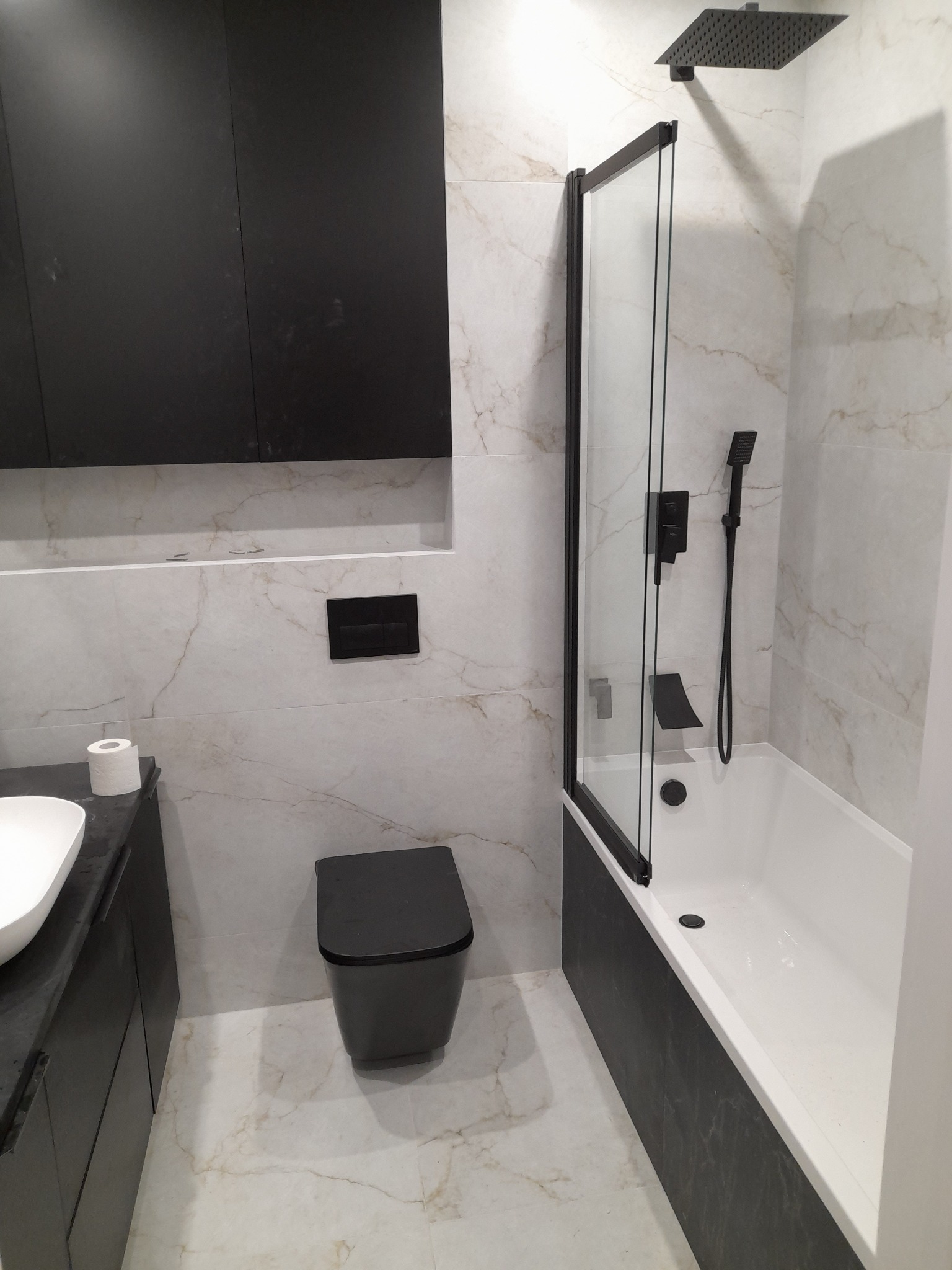 Nowoczesna łazienka z czarną armaturą i białymi marmurkowymi płytkami. Kabina nawannowa z czarną ramą, wisząca toaleta i szafka pod umywalką w ciemnym kolorze.