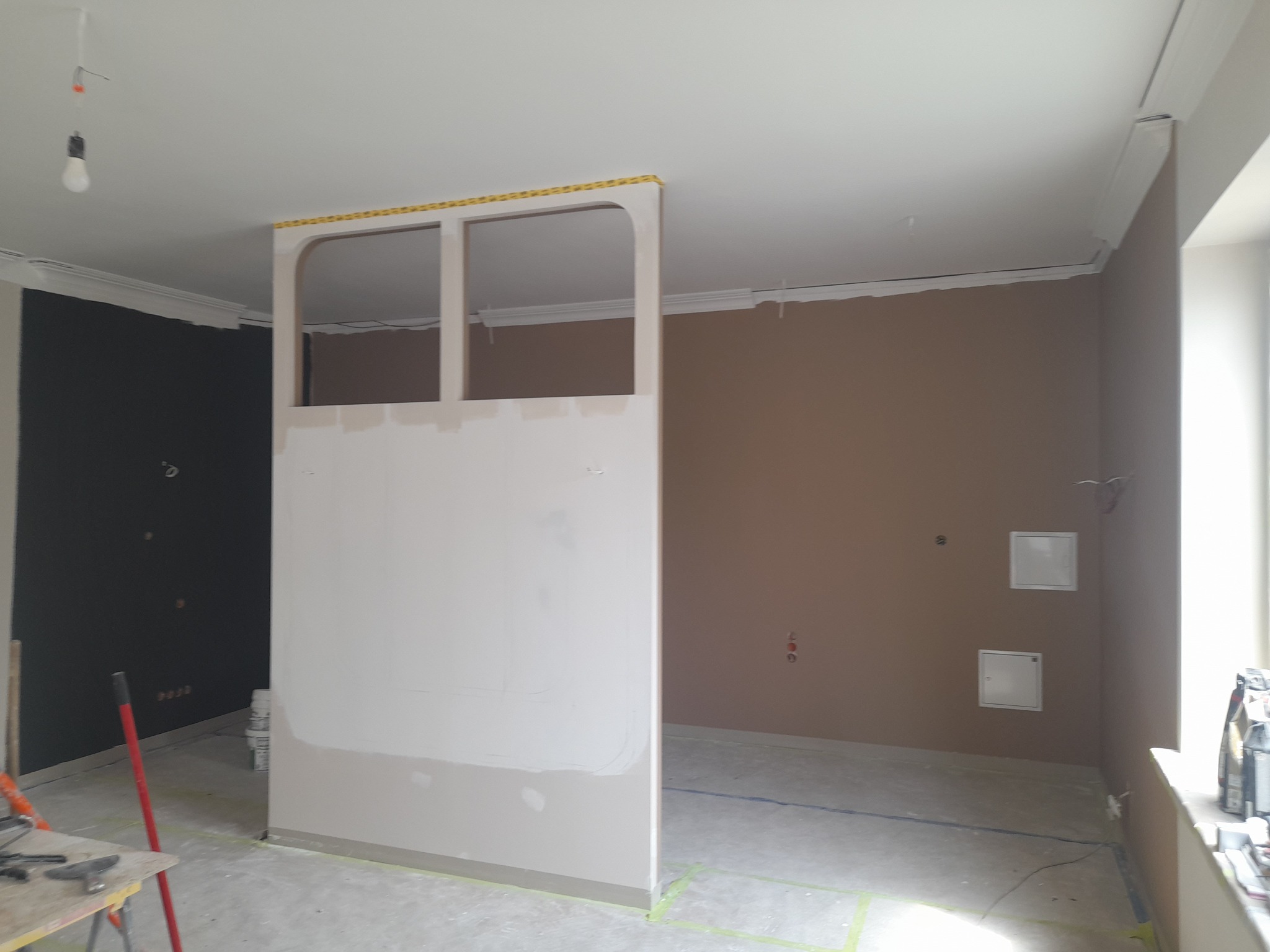 Wnętrze w trakcie prac wykończeniowych: ściany w dwóch kolorach, konstrukcja z płyty gipsowej z otworami, widoczne narzędzia i materiały malarskie.