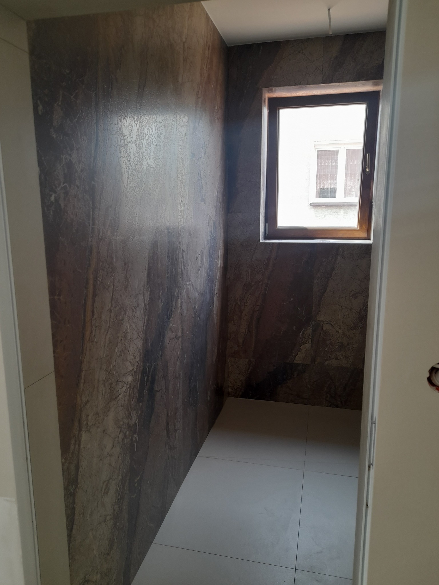 Wykończona łazienka z brązowymi płytkami imitującymi marmur na ścianach i jasnymi płytkami na podłodze. W tle okno z drewnianą ramą. Widok zza otwartych drzwi.