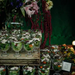 Ustawione rzędem szklane pucharki z sałatką warstwową, dekorowane kwiatami w wazonie i tabliczką informacyjną z nazwą dania na rustykalnym drewnianym stole.