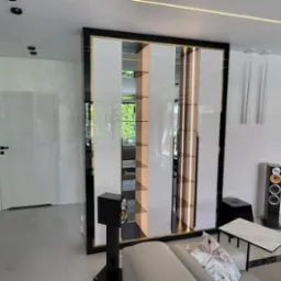 Nowoczesny salon z dużą, podświetlaną szafą wnękową z lustrzanymi elementami, stojącą między ścianą z konsolą a czarnym kominkiem z marmuru.