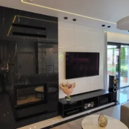 Nowoczesny salon z zabudową meblową: białe szafki, czarny marmurowy kominek z wkładem, telewizor na ścianie, beżowa sofa i okrągły stolik kawowy.