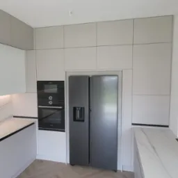 Nowoczesna, jasna kuchnia z szarymi frontami, marmurowym blatem i drewnianą ścianką działową, wyposażona w zabudowany piekarnik, mikrofalówkę i lodówkę side-by-side.