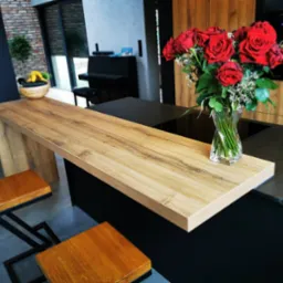 Nowoczesna kuchnia z drewnianym blatem barowym, czarnymi frontami szafek, bukietem czerwonych róż w wazonie i dwoma hokerami z drewnianymi siedziskami na metalowych nogach.