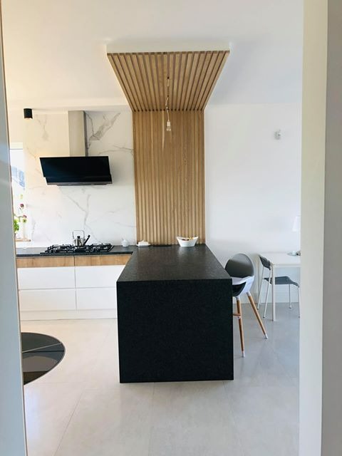 Nowoczesna kuchnia z czarnym wyspowym blatem, białymi szafkami i drewnianą ścianką działową pod sufitem, widok z perspektywy wejścia.