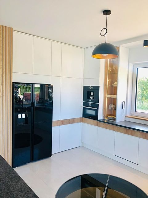 Nowoczesna kuchnia w bieli i drewnie z czarną lodówką side-by-side, zabudowanym ekspresem do kawy i piekarnikiem oraz okrągłym elementem podłogi z czarnego szkła.