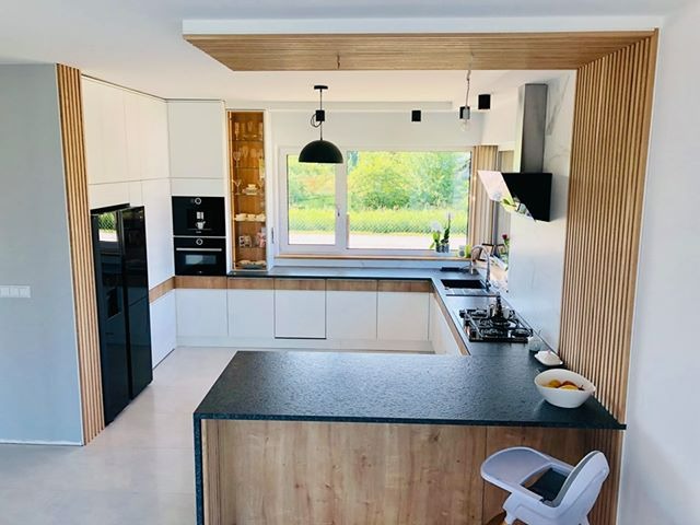 Nowoczesna kuchnia w jasnych barwach z drewnianymi akcentami, widok na okno i ogród, wyspa kuchenna z blatem z ciemnego kamienia.