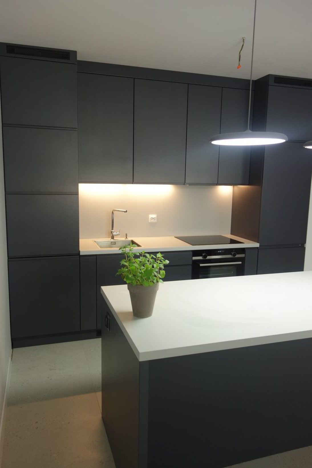 Nowoczesna kuchnia w odcieniach szarości z wyspą i białym blatem, oświetlona ledowym światłem podszafkowym i wiszącą lampą, z doniczką z ziołami na pierwszym planie.