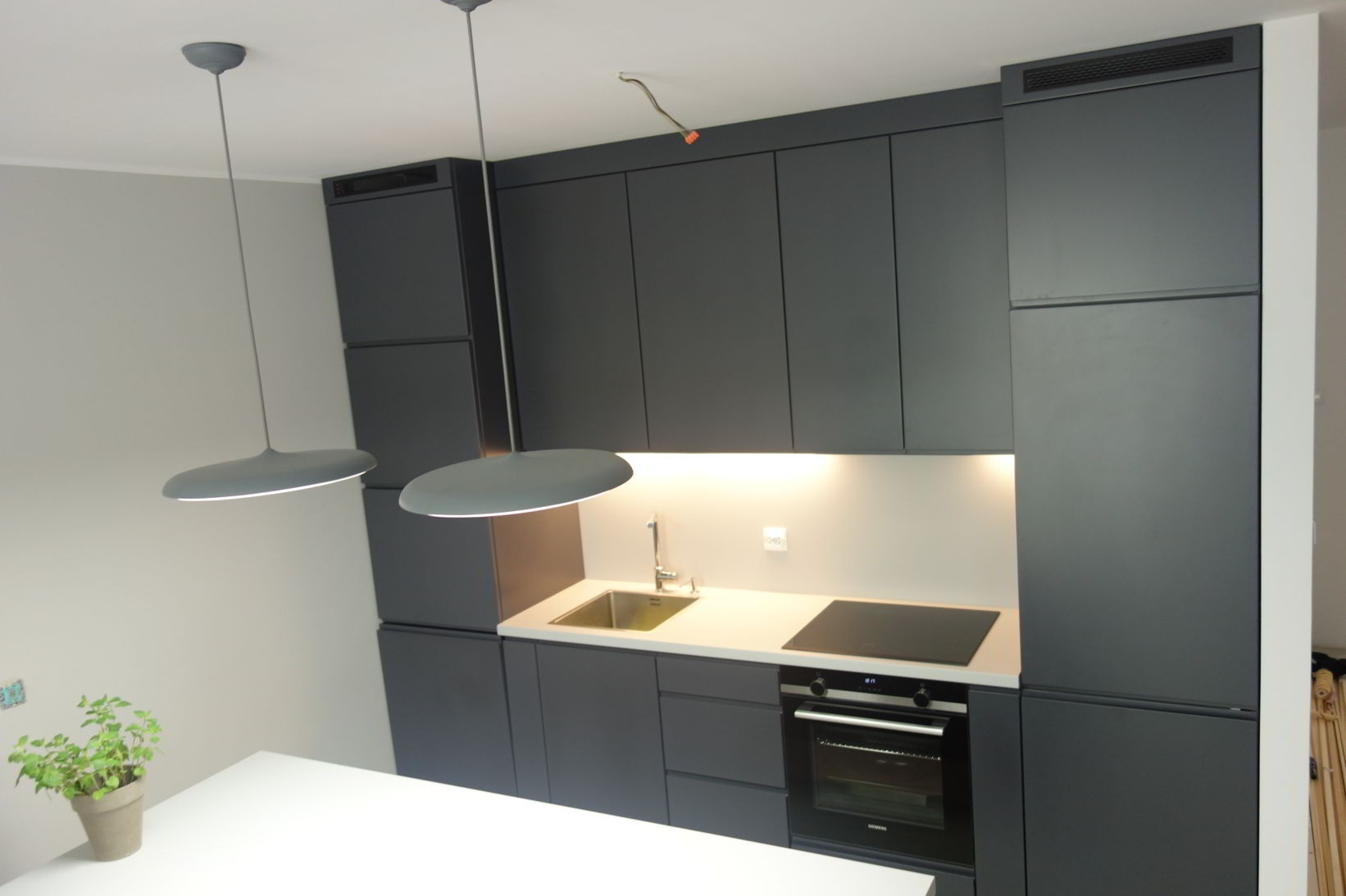 Nowoczesna, antracytowa zabudowa kuchenna z oświetleniem podszafkowym i okrągłymi lampami wiszącymi, widok z góry na blat z płytą indukcyjną i zlewozmywakiem.