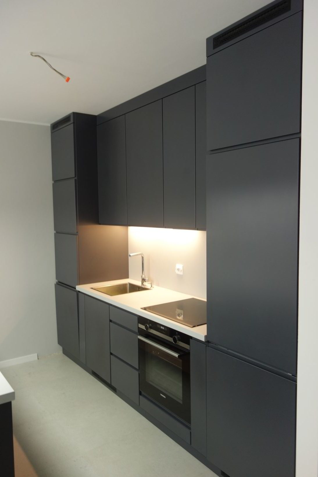Nowoczesna, grafitowa kuchnia na wymiar z oświetleniem LED podszafkowym, minimalistycznym zlewem i płytą indukcyjną, widoczne niedokończone podłączenie elektryczne na suficie.
