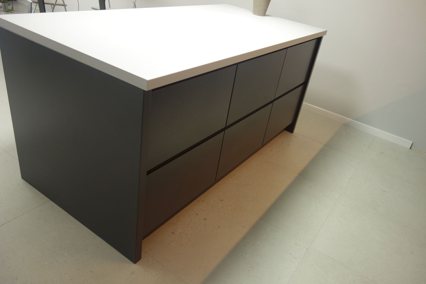 Minimalistyczna wyspa kuchenna z ciemnoszarymi frontami szuflad i białym blatem, widziana z boku, z subtelnym cieniem na podłodze.