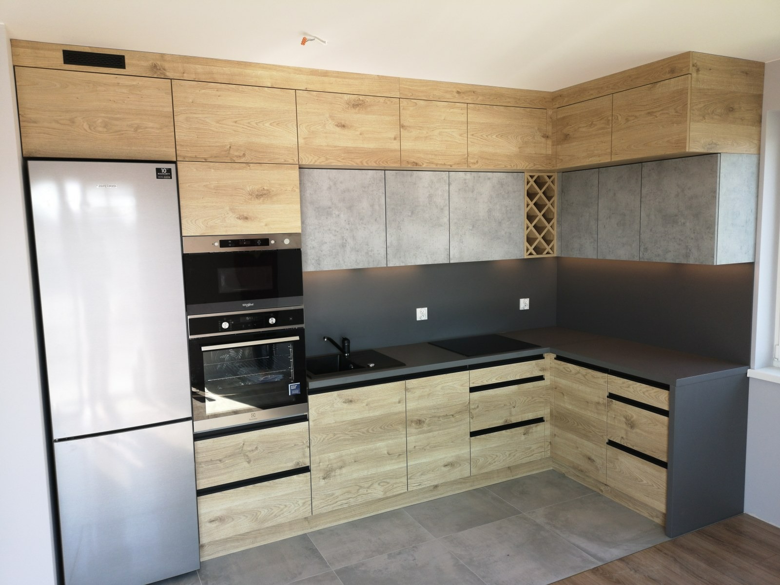 Nowoczesna kuchnia z drewnianymi szafkami, szarymi blatami i sprzętem AGD w zabudowie, widok z narożnika pomieszczenia.