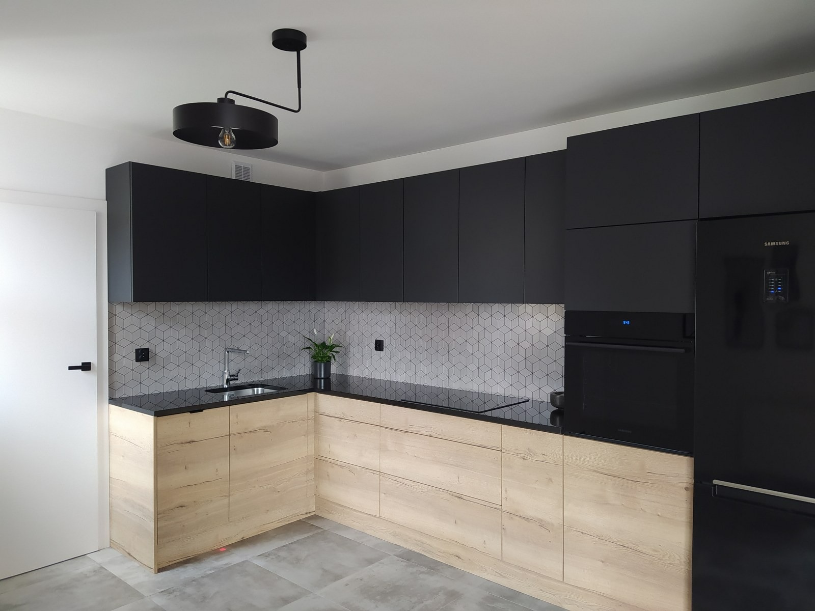 Nowoczesna kuchnia z czarnymi szafkami górnymi i drewnopodobnymi dolnymi, czarny blat, geometryczne płytki na ścianie, czarna lampa wisząca i sprzęt AGD Samsung.