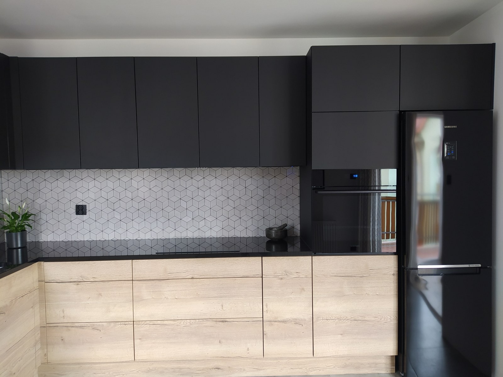 Nowoczesna kuchnia z czarnymi górnymi szafkami i drewnopodobnymi dolnymi szafkami, geometryczna mozaika na ścianie, czarny blat i sprzęt AGD.