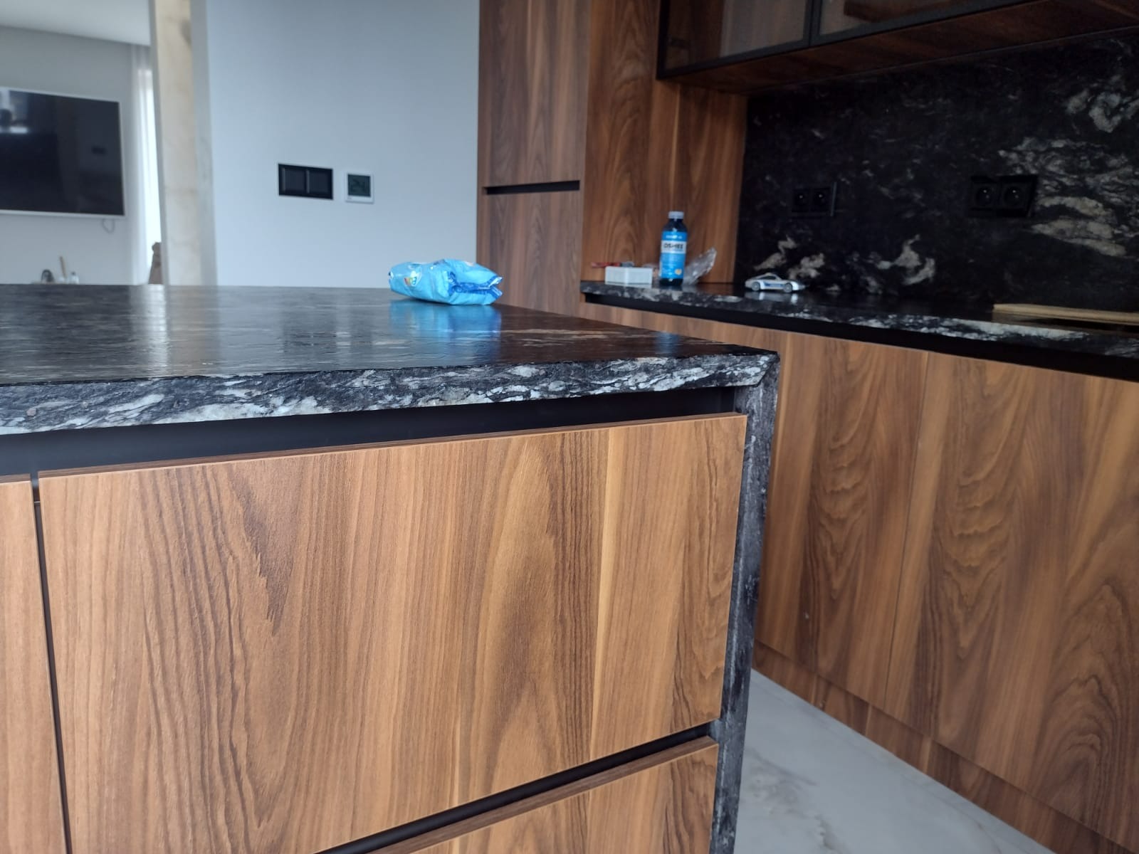 Wyspa kuchenna z blatem z ciemnego granitu i drewnianymi frontami, widoczne gniazdka elektryczne w tle, niebieska ścierka na blacie.