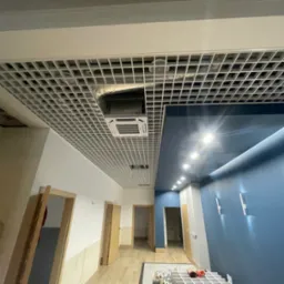 Sufit rastrowy z widoczną instalacją klimatyzacji i oświetleniem w nowocześnie wykończonym wnętrzu z niebieską ścianą i drewnianymi drzwiami.