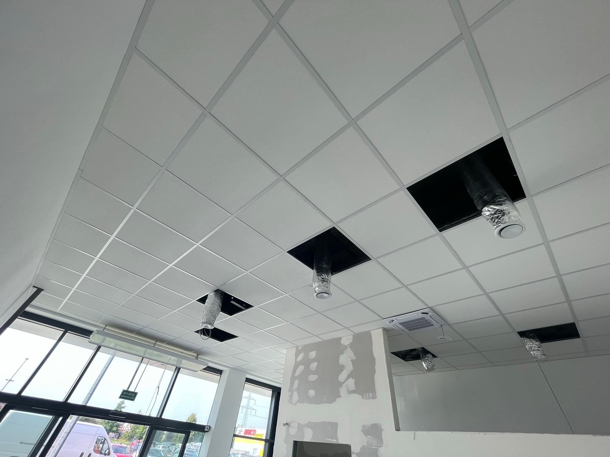 Sufit podwieszany kasetonowy z widocznymi otworami na instalacje wentylacyjne w trakcie montażu, widoczna ściana z niedokończonymi pracami przygotowawczymi pod malowanie.