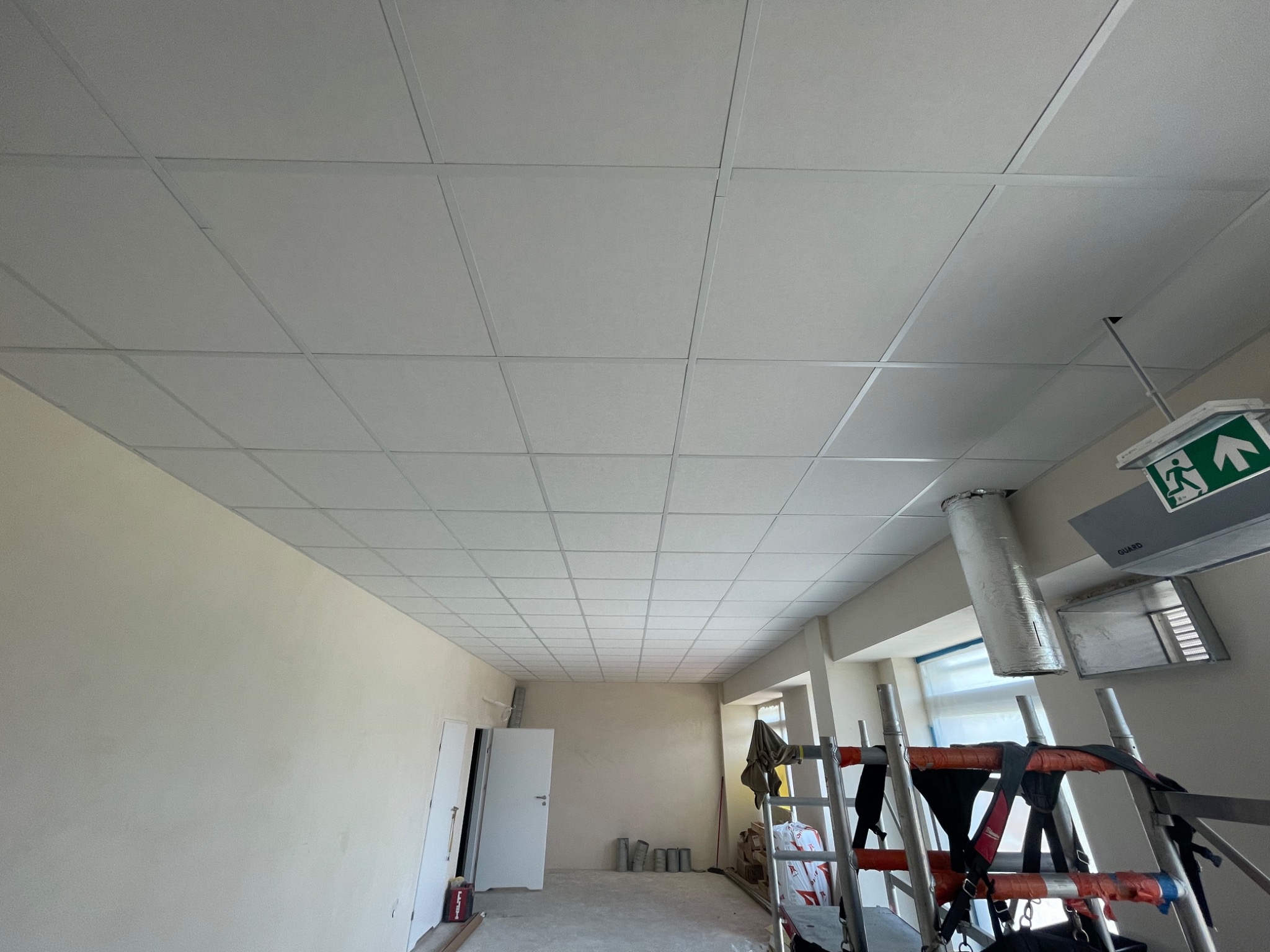 Montaż sufitu podwieszanego kasetonowego w pomieszczeniu biurowym, widoczne rusztowanie, wentylacja i elementy oświetlenia awaryjnego.