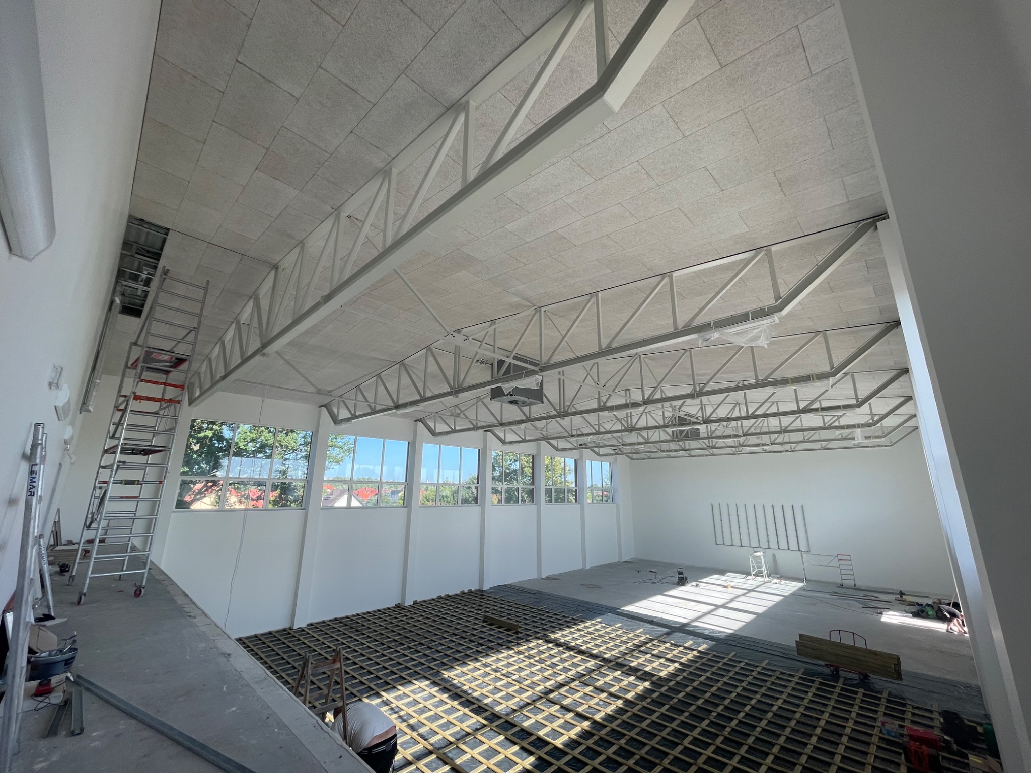 Montaż sufitu z płyt w dużym pomieszczeniu z metalową konstrukcją nośną i widoczną instalacją elektryczną, widok z góry na podłogę z kratownicą i osobę pracującą w rogu.