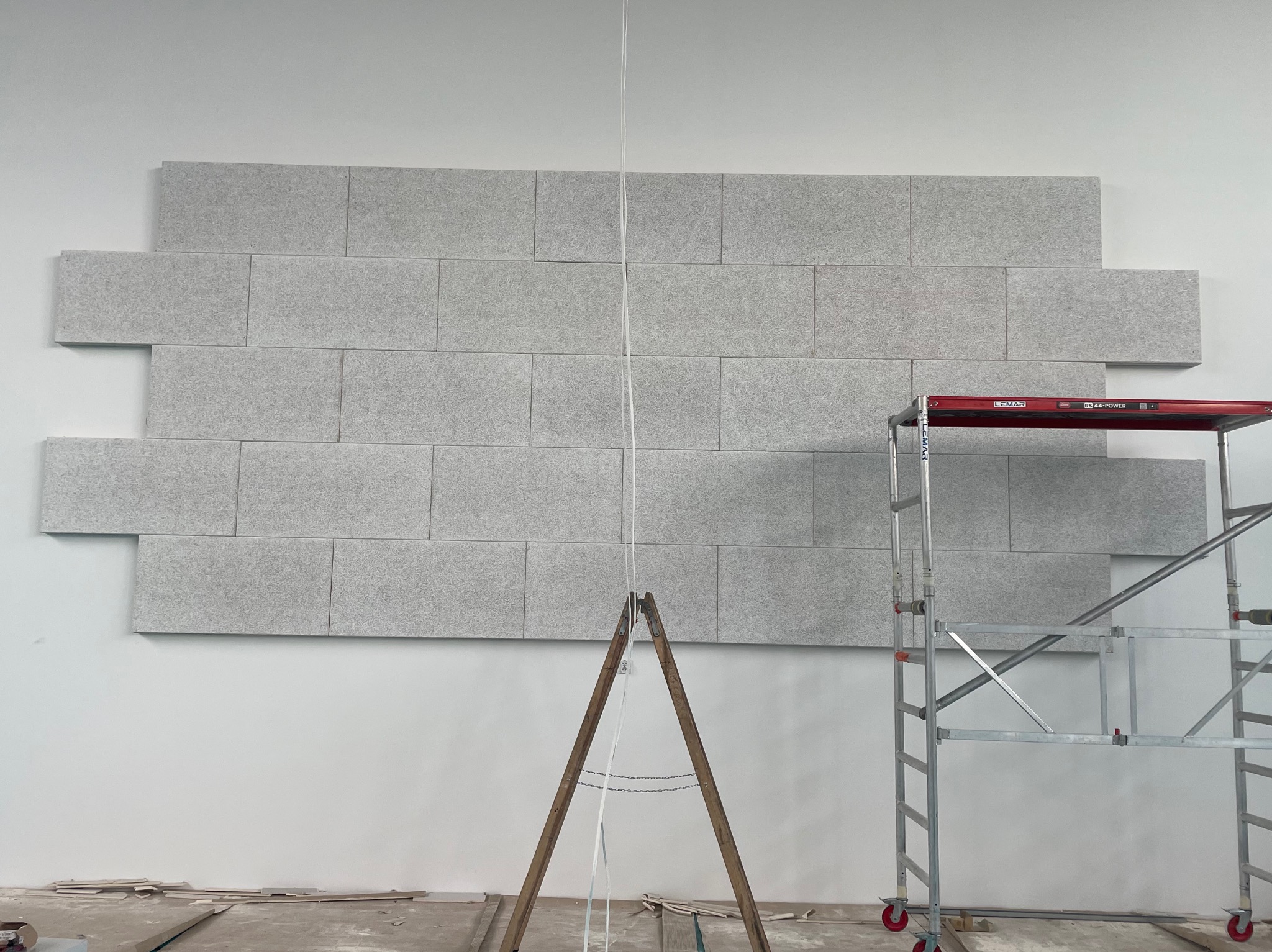 Montaż paneli dekoracyjnych o szarej, teksturowanej powierzchni na białej ścianie, widoczne rusztowanie i drewniany kozioł, fragment podłogi z resztkami materiałów budowlanych.