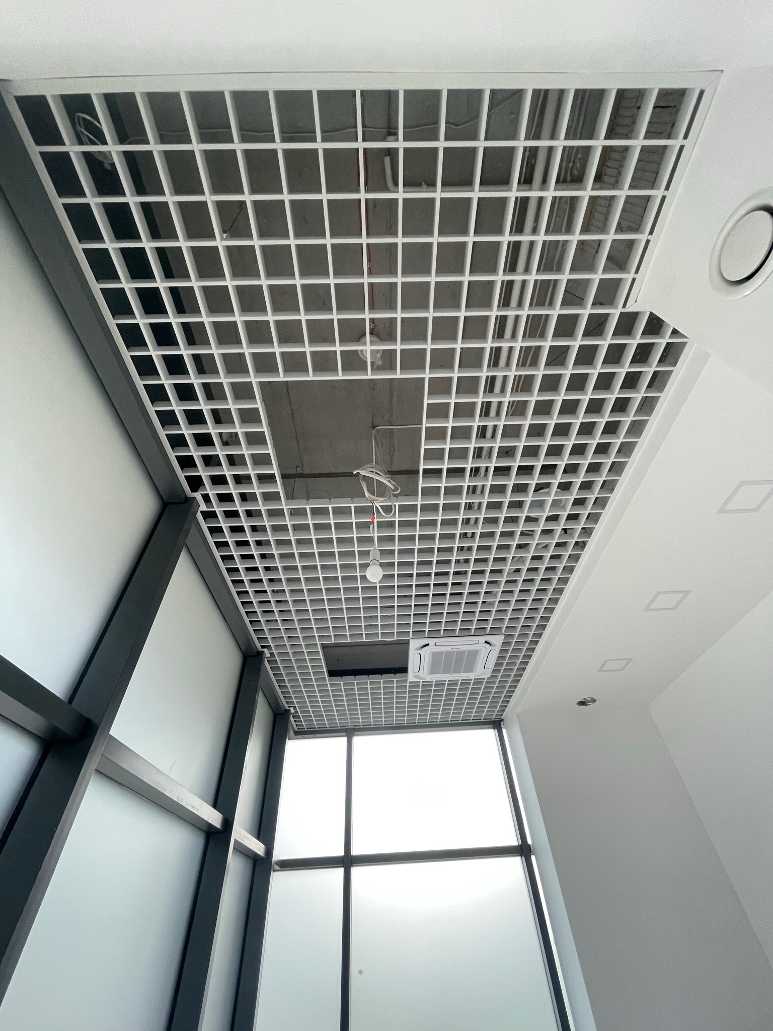 Widok z dołu na biały, rastrowy sufit podwieszany z widoczną instalacją elektryczną i klimatyzacją w przestrzeni między stropem a sufitem, duże okno w tle.