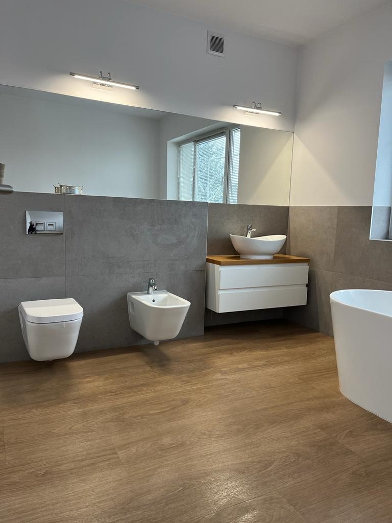 Nowoczesna łazienka z drewnianą podłogą, szarą płytką ścienną, białym umywalką nablatową, bidetem i toaletą w minimalistycznym stylu, oświetlona podłużnymi lampami nad lustrem.