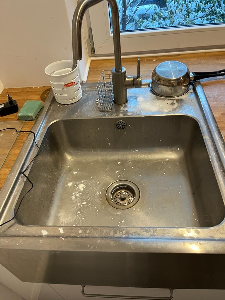 Metalowy zlew kuchenny z resztkami białego proszku czyszczącego, widoczny kran, ociekacz, garnek i akcesoria do mycia naczyń.