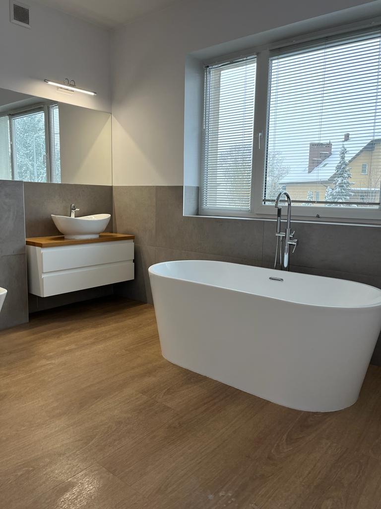Nowoczesna łazienka z wolnostojącą wanną, umywalką nablatową na szafce z drewnianym blatem, szare płytki na ścianach i drewnopodobna podłoga, widok z okna na zaśnieżony ogród.