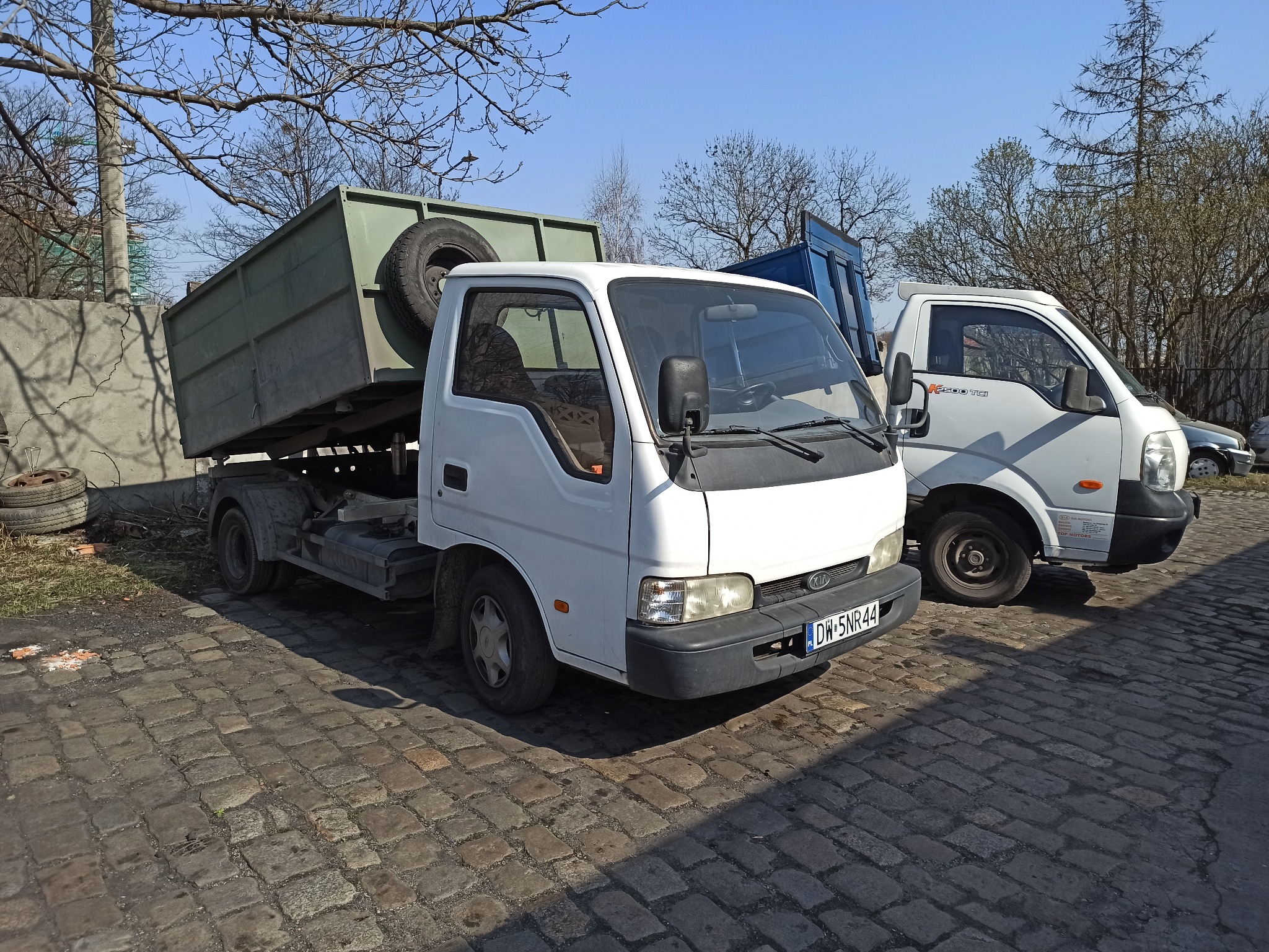 Dwa białe samochody dostawcze Kia, jeden z podniesioną zieloną skrzynią ładunkową, stoją na brukowanym placu w słoneczny dzień.