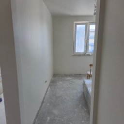 Spontanbud - Wnętrze po remoncie, widok przez futrynę: świeżo pomalowane ściany na biało, surowa podłoga, okno z osłoniętą szybą, krzesło i materiały budowlane w rogu.