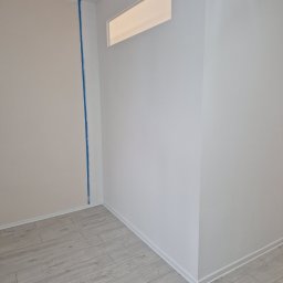 Spontanbud - Wnętrze po remoncie: jasne panele podłogowe, ściany w odcieniach bieli i beżu, z niebieską taśmą malarską w rogu. Nowoczesne wykończenie.