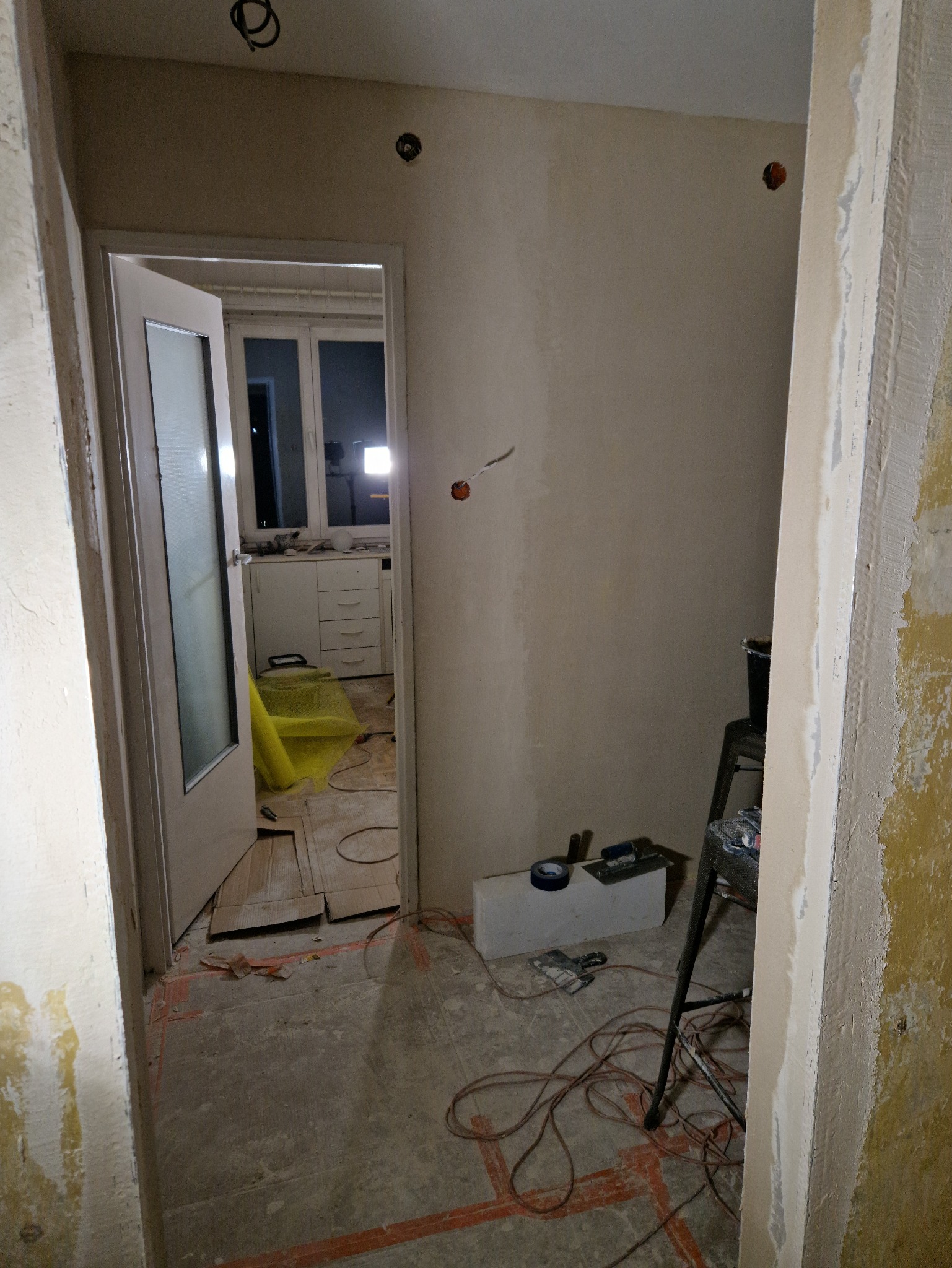 Remont mieszkania: widok na pokój przez otwarte drzwi, ściany w trakcie szpachlowania, widoczne narzędzia i kable na podłodze, w tle kuchnia.