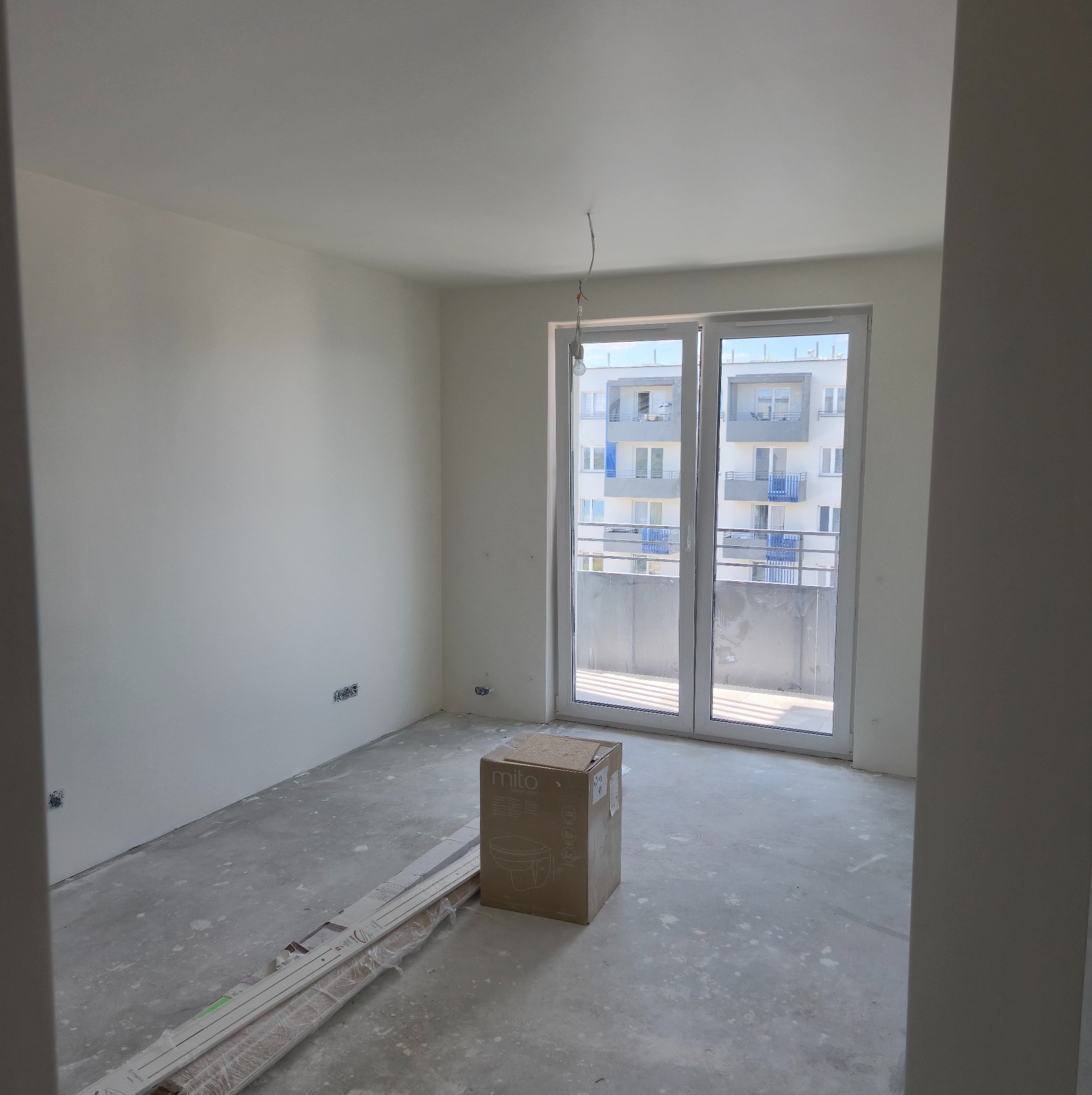 Białe, surowe wnętrze mieszkania w bloku przed wykończeniem: gładkie ściany, okno balkonowe, podłoga wylewka, karton z sanitariatem i panele podłogowe.