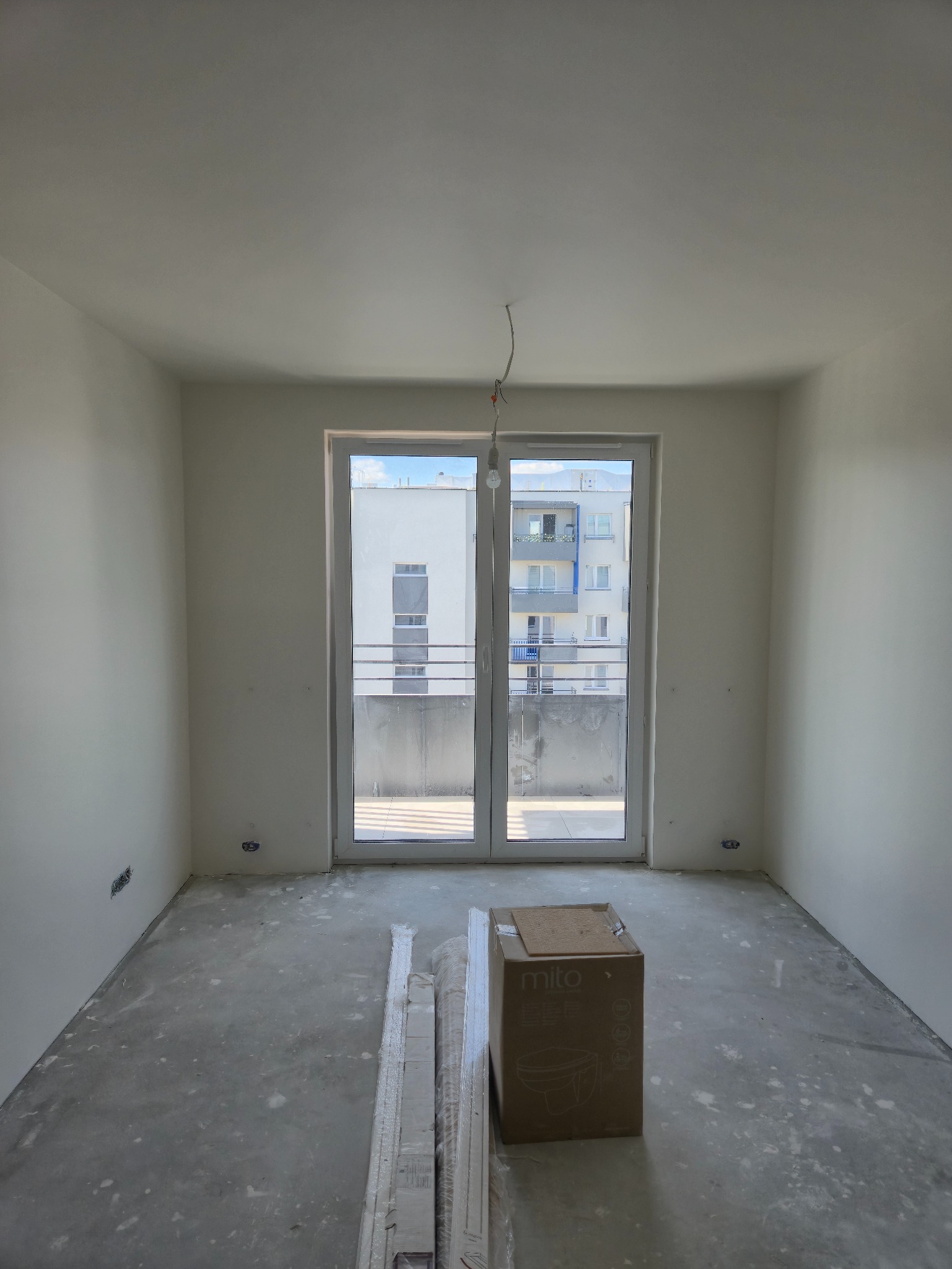Pokój w stanie deweloperskim z białymi ścianami, oknem balkonowym, kartonem i listwami przypodłogowymi na podłodze. Widok na budynek z balkonami.