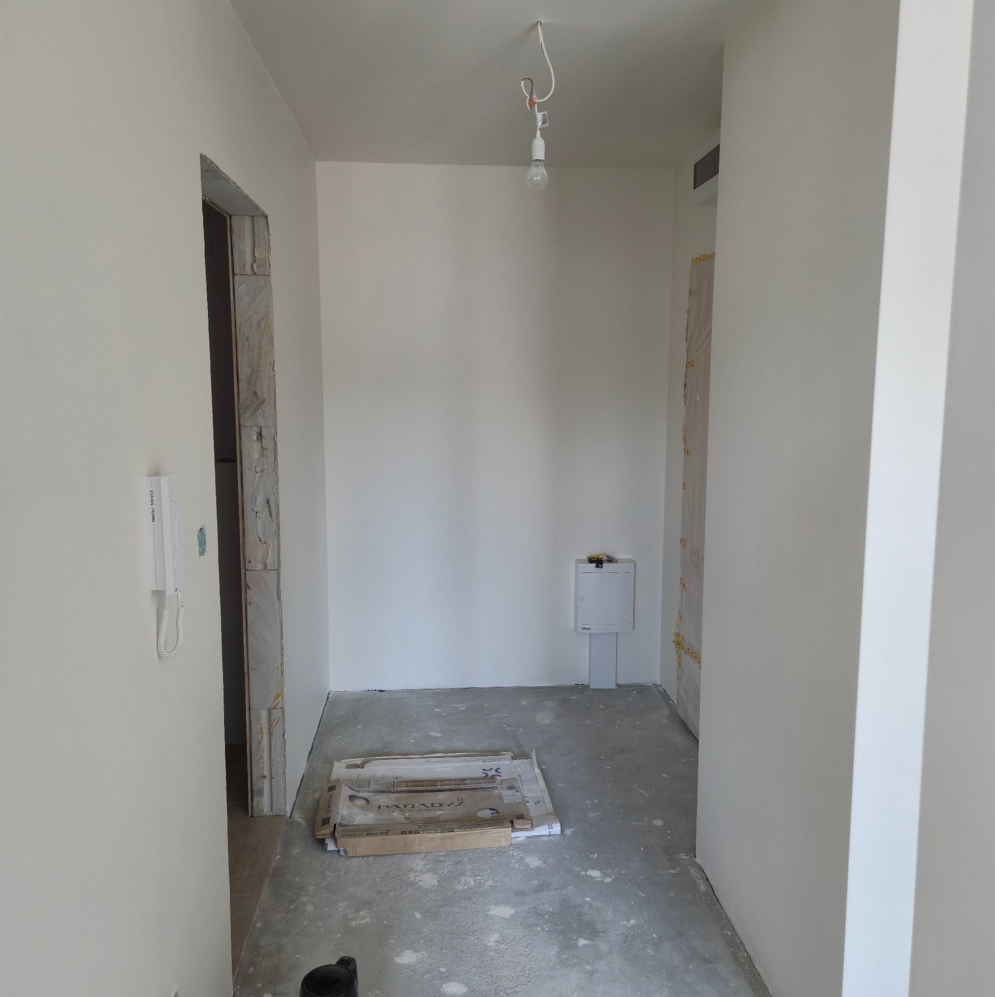 Wnętrze po remoncie: świeżo pomalowane białe ściany, surowy beton na podłodze, kartony z płytkami, widoczne ościeżnice drzwiowe przed montażem.