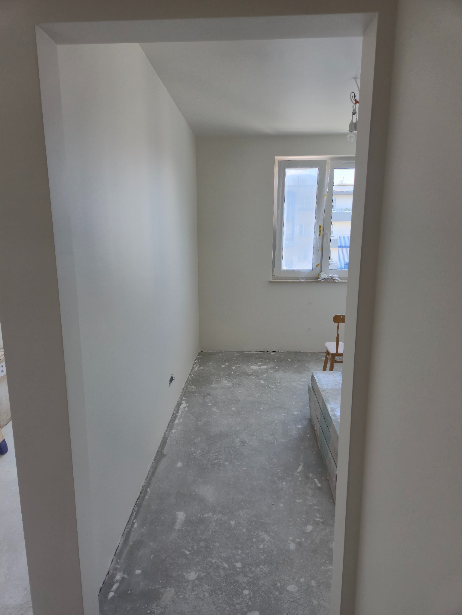 Wnętrze po remoncie, widok przez futrynę: świeżo pomalowane ściany na biało, surowa podłoga, okno z osłoniętą szybą, krzesło i materiały budowlane w rogu.