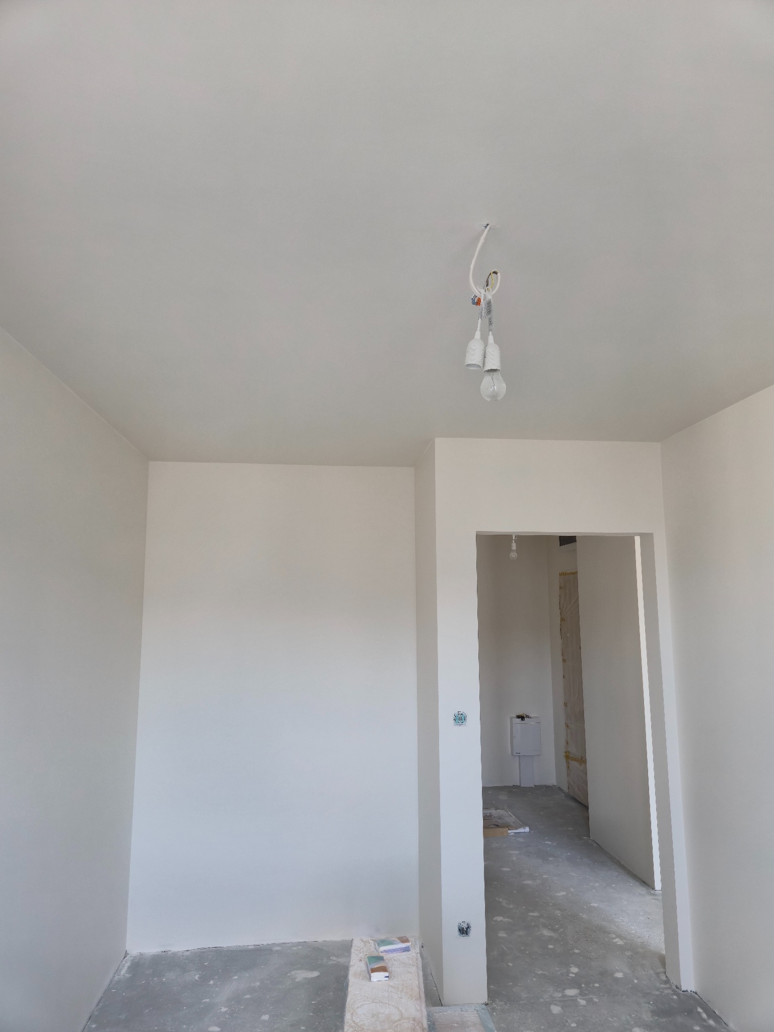 Wnętrze po gładziowaniu w Krakowie. Widoczne świeżo wygładzone, białe ściany, sufit i przygotowane podłoże. W tle widoczny otwór drzwiowy i wisząca żarówka.