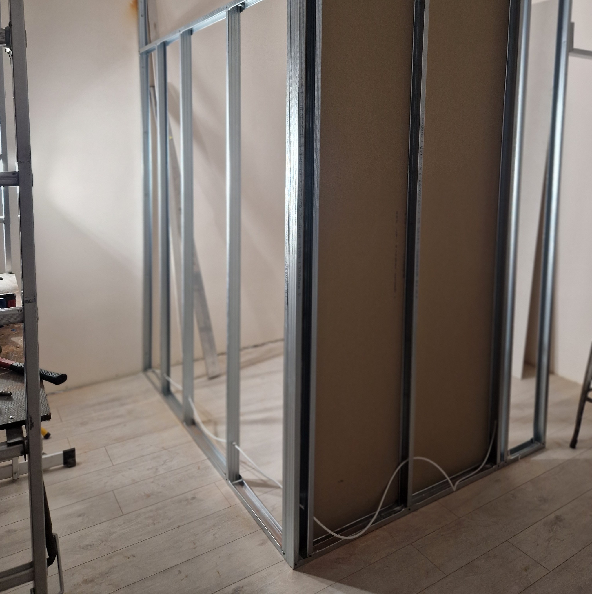 Konstrukcja ścianki działowej z płyt karton-gips na metalowym stelażu, widoczne przewody elektryczne, podłoga z paneli, fragment drabiny.