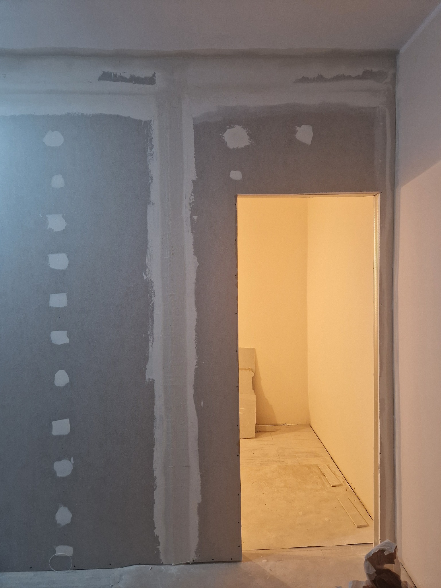 Ściana z płyt gipsowo-kartonowych w trakcie szpachlowania, widoczne łączenia płyt i otwór drzwiowy. W tle jasne pomieszczenie w trakcie prac wykończeniowych.