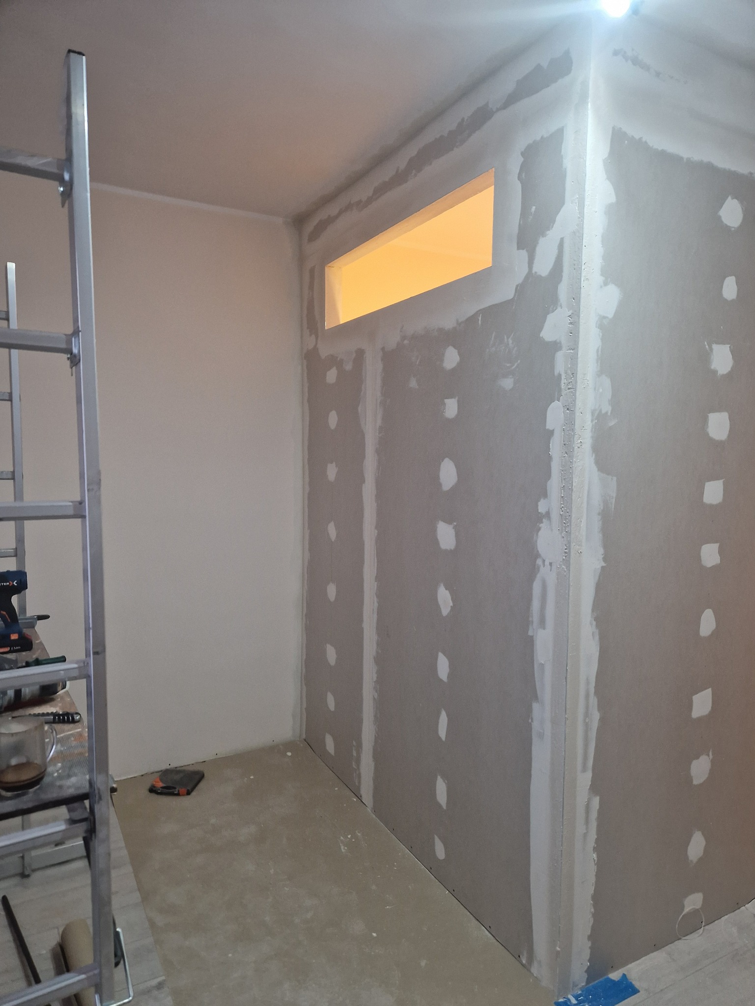 Wnętrze w trakcie prac wykończeniowych: ściany z płyt gipsowo-kartonowych, widoczne łączenia i szpachlowane miejsca po wkrętach, drabina w rogu pomieszczenia.