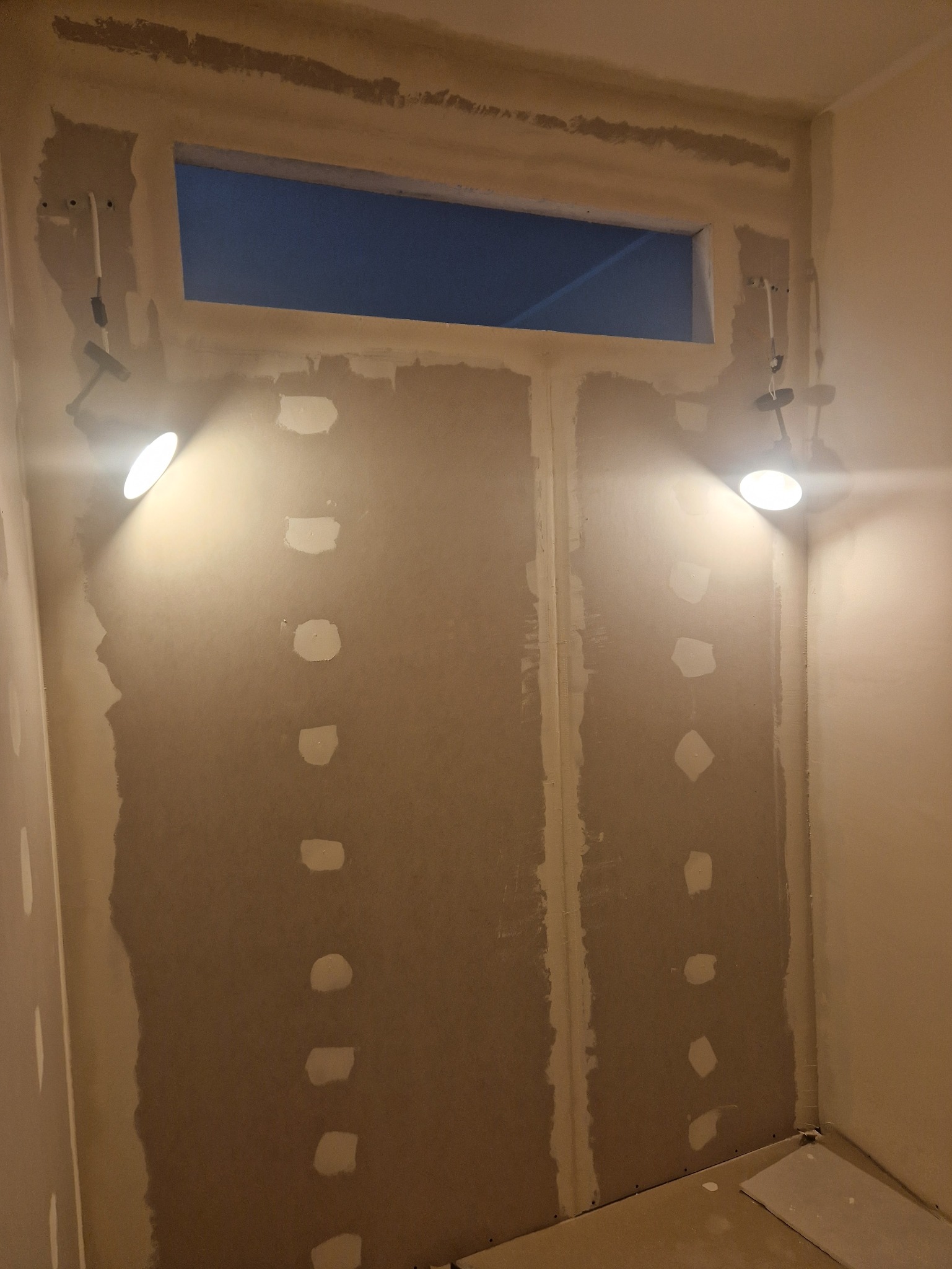 Ściana z płyty karton-gips przed szpachlowaniem, oświetlona dwoma lampami halogenowymi. Widoczne łączenia płyt i miejsca po wkrętach wypełnione gipsem.