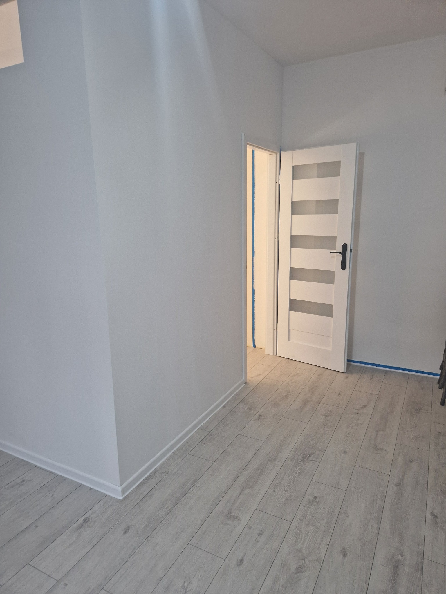 Jasne wnętrze po remoncie: białe ściany, panele podłogowe. Nowe, białe drzwi z przeszkleniami. Taśma malarska zabezpiecza krawędzie. Minimalistyczny styl.