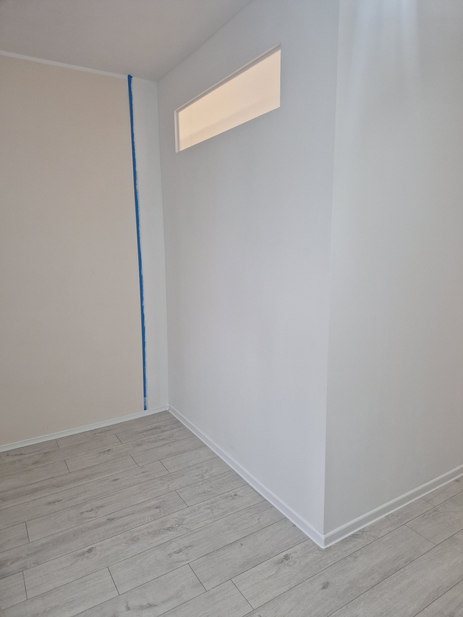 Wnętrze po remoncie: jasne panele podłogowe, ściany w odcieniach bieli i beżu, z niebieską taśmą malarską w rogu. Nowoczesne wykończenie.