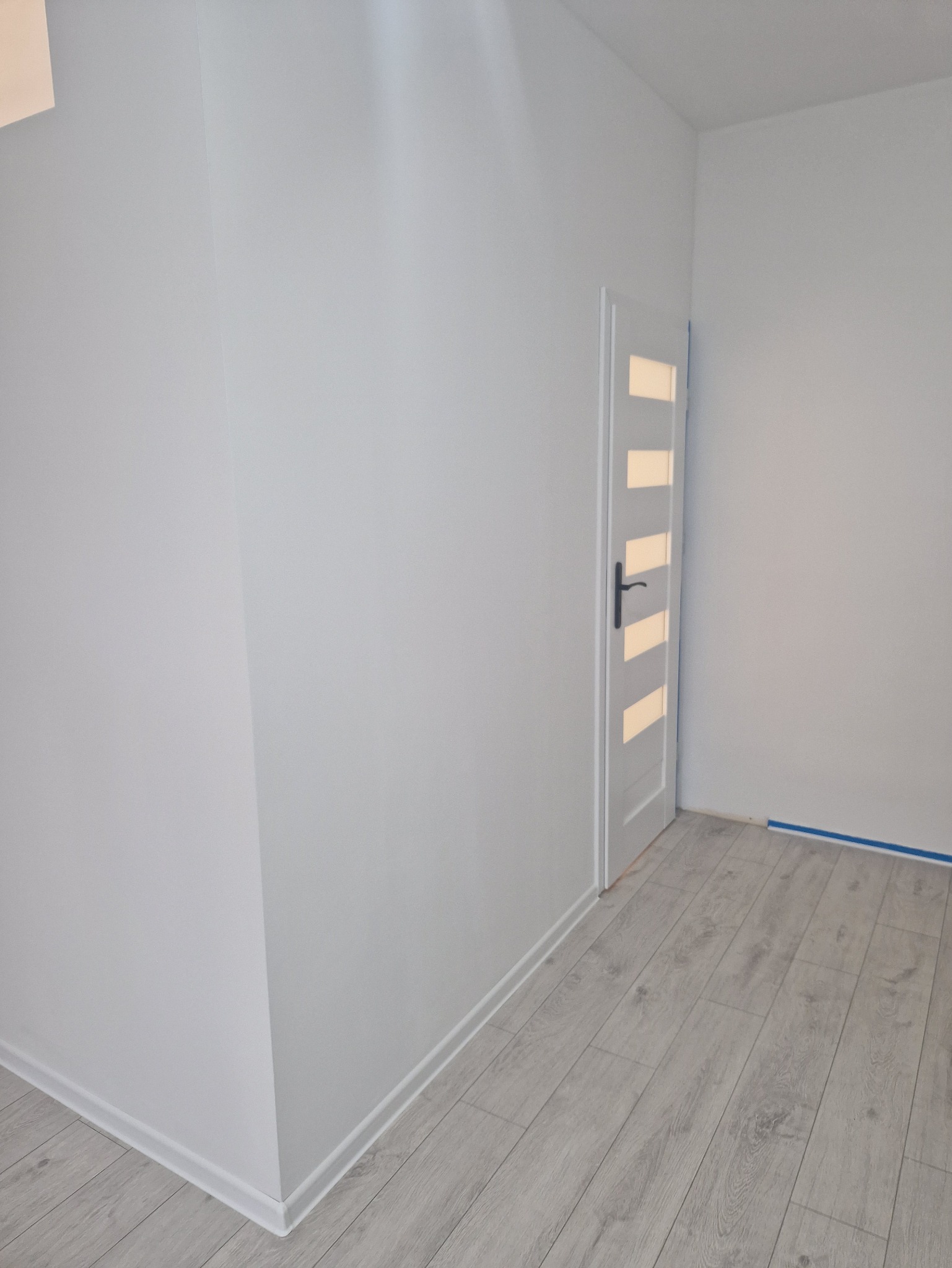 Jasne wnętrze po remoncie: białe ściany z listwami przypodłogowymi, panele podłogowe w odcieniach szarości i białe drzwi z przeszkleniami.