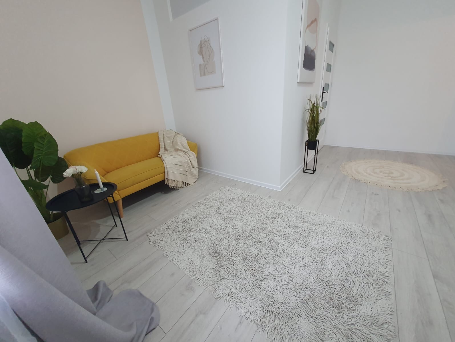 Jasne wnętrze po remoncie: żółta sofa z pledem, stolik z kwiatami, puszysty dywan i drewniana podłoga. Minimalistyczne obrazy na ścianach dopełniają aranżację.
