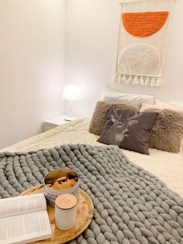 Przytulna sypialnia z dekoracyjnym gobelinem, pledem z grubej wełny i poduszkami. Na łóżku taca z książką i ciasteczkami. Ciepłe oświetlenie podkreśla domowy charakter.