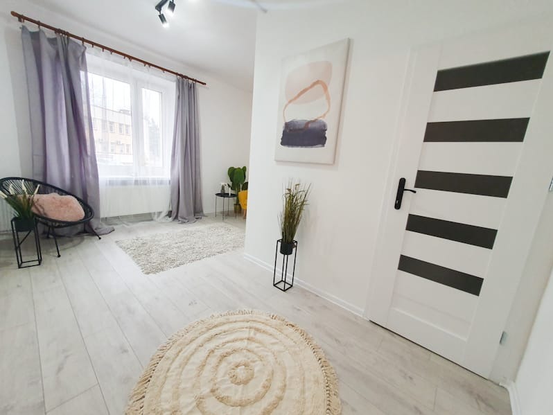 Jasne wnętrze pokoju po remoncie: białe ściany, panele podłogowe, białe drzwi z czarnymi pasami, okno z zasłonami i dekoracje w stylu minimalistycznym.