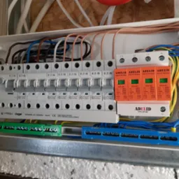 Rozdzielnica elektryczna z zabezpieczeniami przepięciowymi Adelid, widoczne bezpieczniki Eaton, przewody w kolorach niebieskim, żółtym, brązowym i białym, zamontowana w skrzynce na drewnianym podłożu.