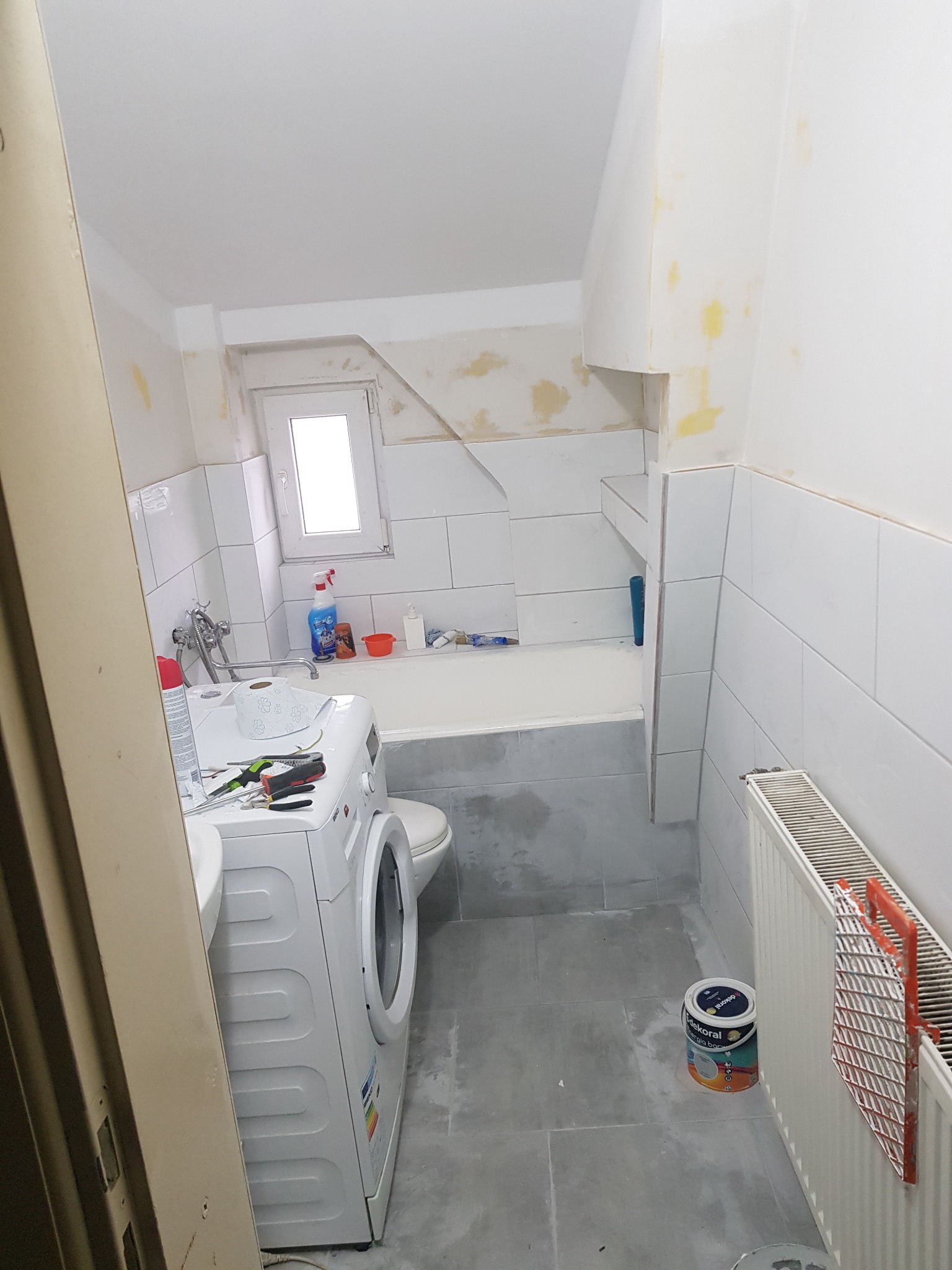 Łazienka w trakcie remontu: widoczne płytki, wanna, umywalka z pralką, okno, grzejnik z wałkiem malarskim i puszka farby Dekoral na podłodze.